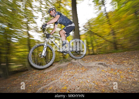 Mountainbiker in Vienna Woods rides fast along trail through the autumn forest, jumps over big root. Side view of athletic cyclist with motion blur. Stock Photo