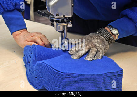 A seamstress uses a vertical knife to cut out a pile of fabric for jackets in a commercial sewing room. Stock Photo