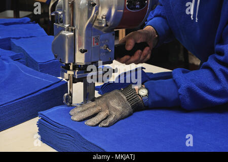 A seamstress uses a vertical knife to cut out a pile of fabric for jackets in a commercial sewing room. Stock Photo