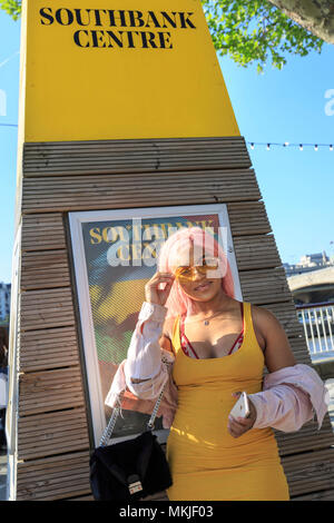 South Bank, London, 7th May 2018. A young lady in a striking yellow summer dress and yellow glasses happily poses in the sunshine, colour-matching a yellow 'Southbank Centre' sign. Tourists and Londoners enjoy the sunny weather on the South Bank of London by the river Thames, with record breaking temperatures of up to 28 degrees. Credit: Imageplotter News and Sports/Alamy Live News Stock Photo