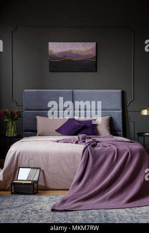 Purple blanket on the bed in dark bedroom interior with a painting on grey wall Stock Photo
