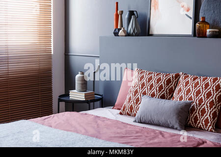 Close-up of patterned pillows lying on a soft bed in bedroom interior with black walls, decorative vases and brown blinds. Real photo Stock Photo