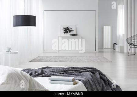 Books on white bed in spacious, minimal bedroom interior with black lamp and poster Stock Photo
