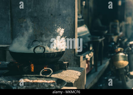 Street food in India cooking in fatiscent big pan or wok in a small street food stall. White smoke coming out from the pan, teal toned image. Stock Photo