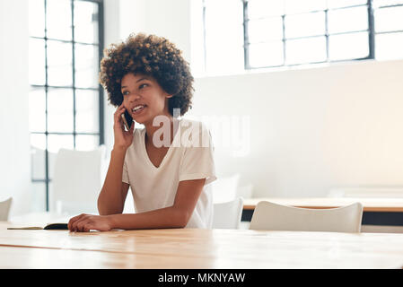 Casually dressed young African female student with an afro smiling and talking on her cellphone while sitting alone at a table on campus Stock Photo