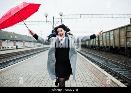 Brunette girl in gray coat with red umbrella in railway station. Stock Photo