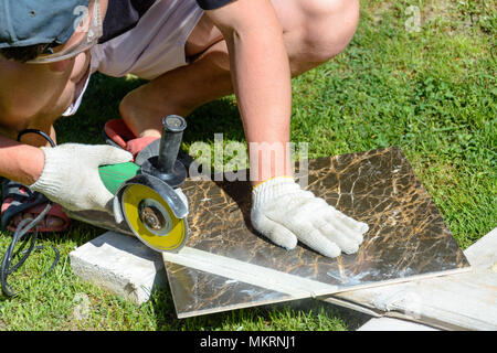 The working tiler cuts the tile with a corner grinder on the floor, movement in the frame, dusty dirty work Stock Photo
