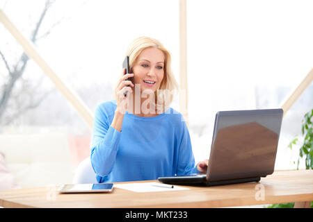 Smiling middle aged businesswoman making call and using laptop while sitting at desk and working. Home office. Stock Photo