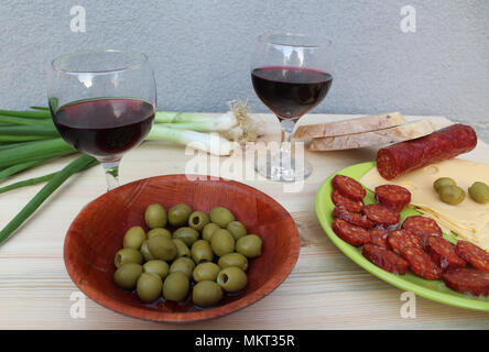 Garden party in Mediterranean style - two glasses of red wine, green olives in bamboo bowl and sliced sausage, cheese and bread on a green plate Stock Photo