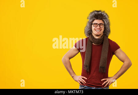 Laughing stylish man in t-shirt and winter scarf with furry hat smiling at camera standing on yellow background. Stock Photo