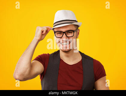 Handsome man in stylish hat and glasses standing on yellow background smiling at camera and touching hat. Stock Photo