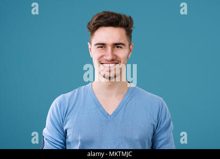 Young casual man with trendy hairstyle wearing blue pullover and looking with smile at camera on blue. Stock Photo