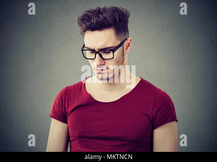 Handsome casual man in eyeglasses looking down with feeling of sadness standing on gray background. Stock Photo