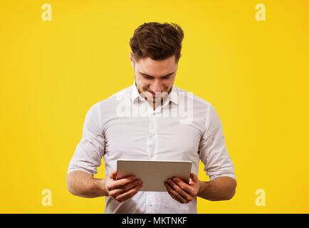 Young handsome man in white shirt looking happy while surfing new tablet on yellow background. Stock Photo