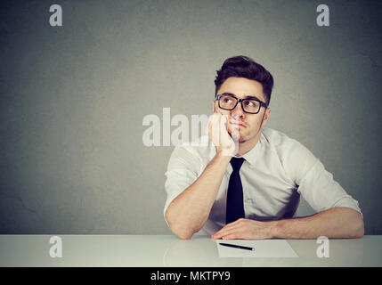 Young man in eyeglasses sitting with paper and pen at table and looking away in contemplation. Stock Photo