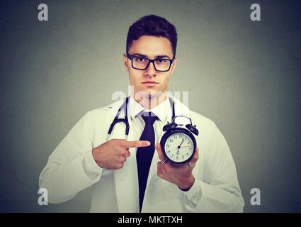 Young handsome man in medical uniform holding and pointing at clock looking confidently at camera. Stock Photo