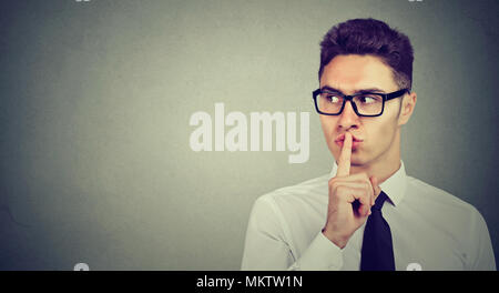 Secret guy. Man saying hush be quiet with finger on lips gesture looking to the side isolated on gray wall background. Stock Photo