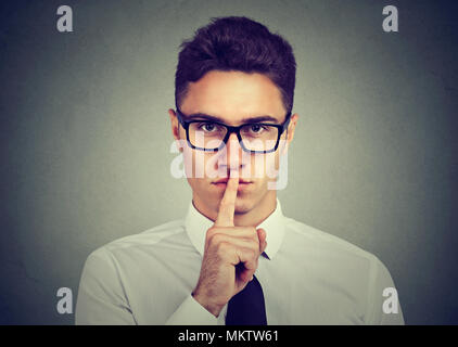 Keep my secret. Businessman holding finger on lips looking at camera standing against gray background Stock Photo