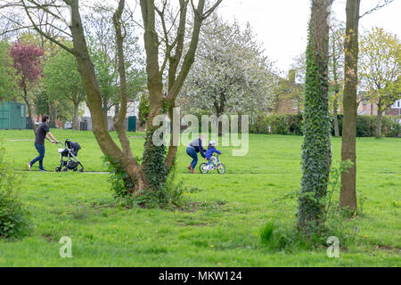 Father pushes a baby in its pram while mother helps their son on a bicycle in Bruche Park, Warrington, Cheshire, England on 29 April 2018 Stock Photo