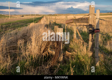 Spring thunderstorm clouds in Saskatchewan with a fencepost, roll of barbed wire, tall grass and a road in the foreground Stock Photo