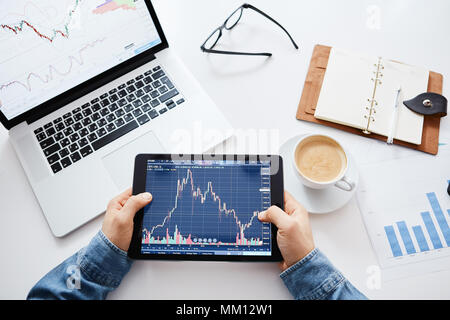 Young trader is sitting in home at table, working on tablet with graphs,diagrams on monitor. Crypto currencies, trader using tablet and computers. Stock Photo