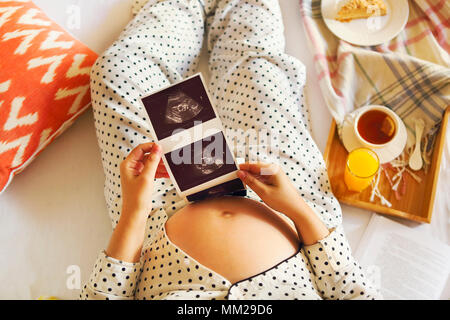Pregnant woman wearing pijama with book, tea, cake relaxing at home. Top view