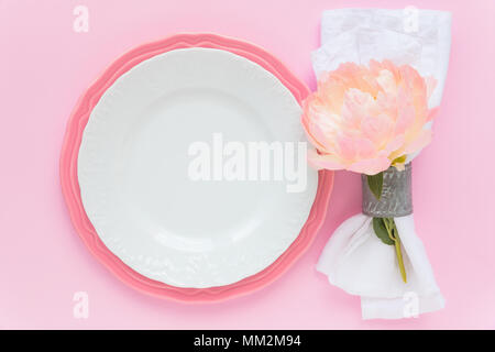 Summer table place setting with white an d pastel pink plates white linen napkin decorated with fresh peony flower on pink background. Overhead view Stock Photo