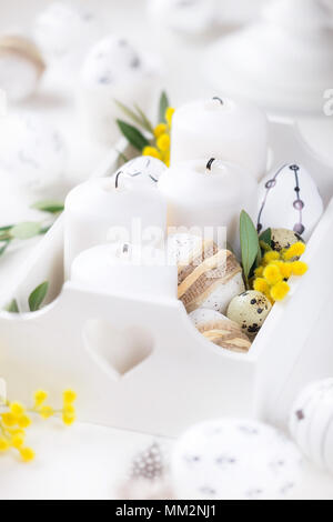 Beautiful Easter composition with white unlit candles in a white wooden box with decorated Easter eggs, olive branches and yellow mimosa flowers as a  Stock Photo