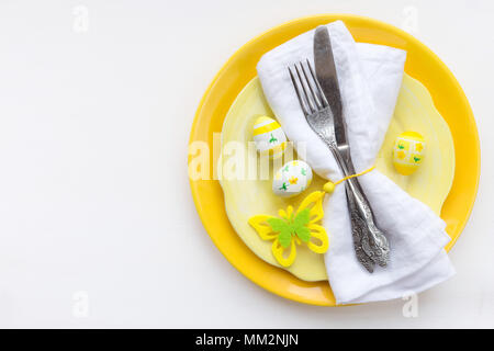 Easter table setting with yellow plates and silverware wrapped in white linen napkin and traditional Easter decorations on white wooden table Stock Photo