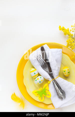 Easter table setting with yellow plates and vintage silverware wrapped in white napkin with traditional Easter decorations on white wooden background. Stock Photo