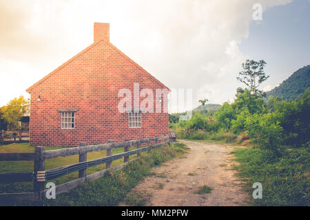 Beautiful landscape image of old brick house surrounded with wooden fence beside dirt road at countryside in rainy day. Stock Photo