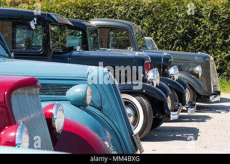 Shiny, polished hot rods parked side by side, outside in sunshine. Collection of old, classic, American cars, taken from side showing front bumpers. Stock Photo