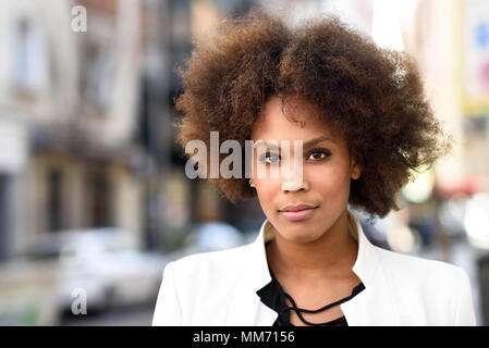 Young black woman with afro hairstyle standing in urban background. Mixed girl wearing white jacket and black dress posing near a brick wall Stock Photo