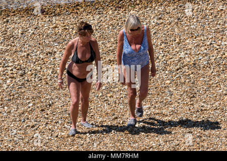 two older ageing women walking on a shingle beach on a hot summers day wearing swimming costumes. retired pensioners in swimsuits walking on pebbles. Stock Photo