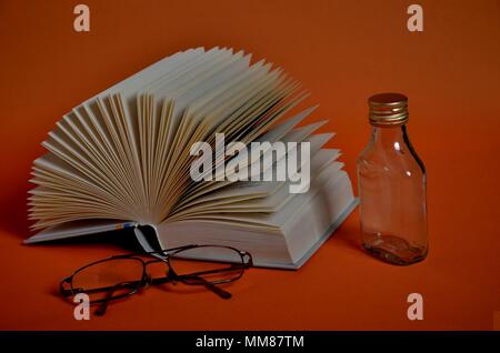 Reading glasses, open book forming a fan and small liquor on orange background Stock Photo