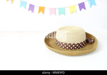 Straw hat on white table with colorful party flags, bunting decoration. Greeting card, invitation for summer birthday or Brazilian june party, Festa Junina holiday. Midsummer celebration. Stock Photo