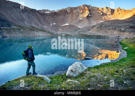 Bearded man in green hat and backpack looking at mountain lake at sunrise Stock Photo