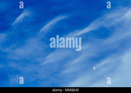 Crescent moon behind high altitude cirrus clouds against a blue sky Stock Photo