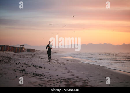 Full length shot of fit male runner running on the beach in morning. Fitness man jogging on the sea shore. Stock Photo