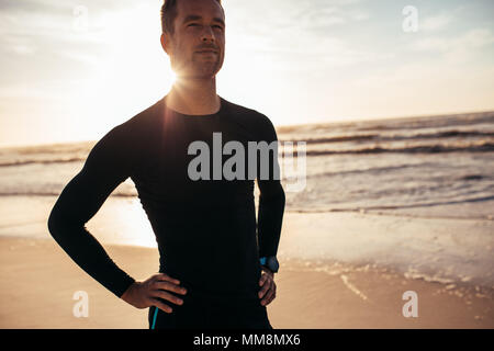 Portrait of confident male athlete standing on the beach. Man in sportswear along the sea shore in morning. Stock Photo