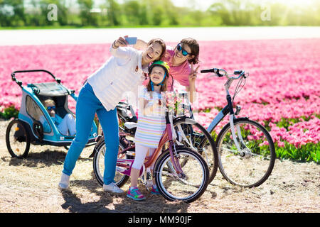 Dutch family riding bicycle in tulip flower fields in Netherlands. Mother and kids taking selfie picture with mobile phone camera on bikes at blooming Stock Photo