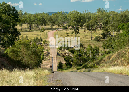 Steep road leading down to narrow low bridge across Bowen River & through landscape of trees & golden grasses under blue sky in Queensland Australia Stock Photo
