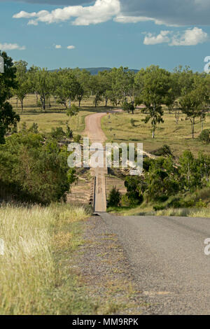 Steep road leading down to narrow low bridge across Bowen River & through landscape of trees & golden grasses under blue sky in Queensland Australia Stock Photo
