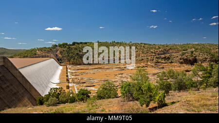 Burdekin Falls dam with high spillway overflowing and coffee coloured water flowing into rocky river bordered by wooded hills under blue sky, Qld Aust Stock Photo