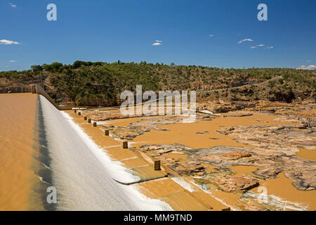 Burdekin Falls dam with high spillway overflowing and coffee coloured water flowing into rocky river bordered by wooded hills under blue sky, Qld Aust Stock Photo