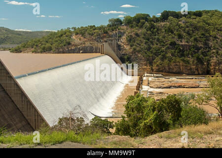 Burdekin Falls dam with high spillway overflowing and coffee coloured water flowing into rocky river bordered by wooded hills under blue sky, Qld Aust Stock Photo