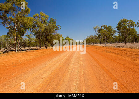 Long straight red dirt road slicing through Australian outback landscape with scattered eucalyptus trees and stretching to distant horizon under blue Stock Photo
