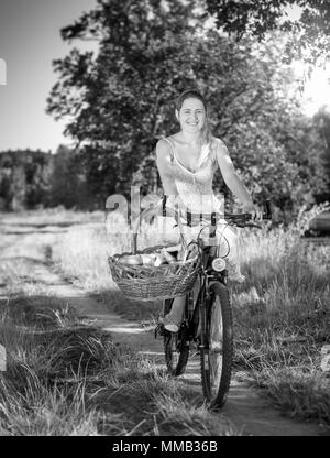 Black and white image of beautiful smiling woman riding on bicycle with big basket for picnic in park Stock Photo