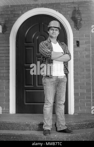 Black and white image of handsome young builder posing in front of door of new house Stock Photo