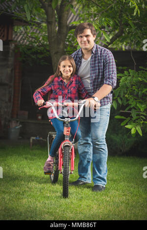 Toned photo of happy young man teaching his daughter how to ride bicycle Stock Photo